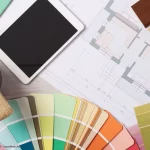Grundriss, Tablet, Farbfächer, Stoffmuster und Pinsel auf einem Schreibtisch veranschaulichen die kreative Raumgestaltung und Materialauswahl beim Interior Design.